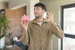 Individual Savings Account: young handsome man feeling happy, showing approval with okay gesture. piggy bank concept